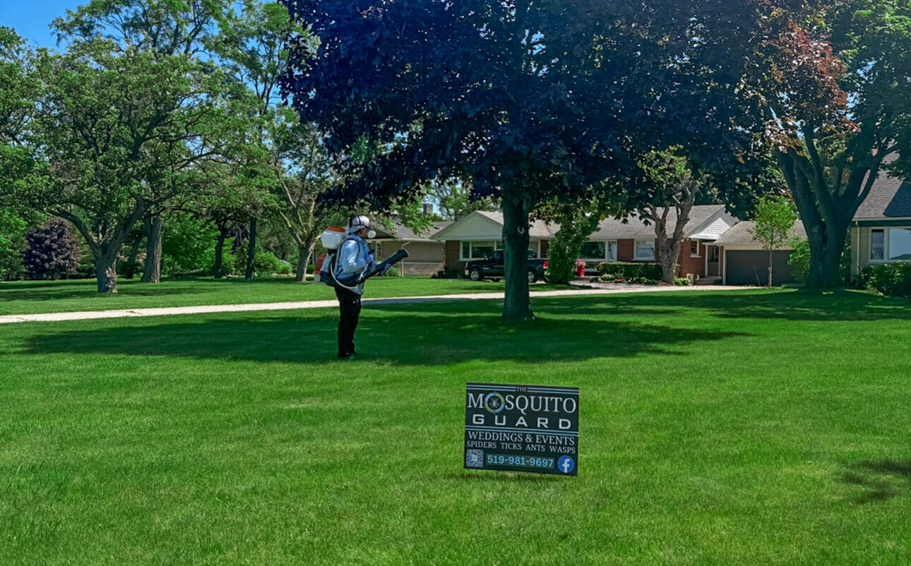 A Mosquito Guard technician in protective gear sprays a residential lawn with pest control treatment on a sunny day. A branded yard sign in the foreground advertises services for weddings, events, and pest control for spiders, ticks, ants, and wasps.