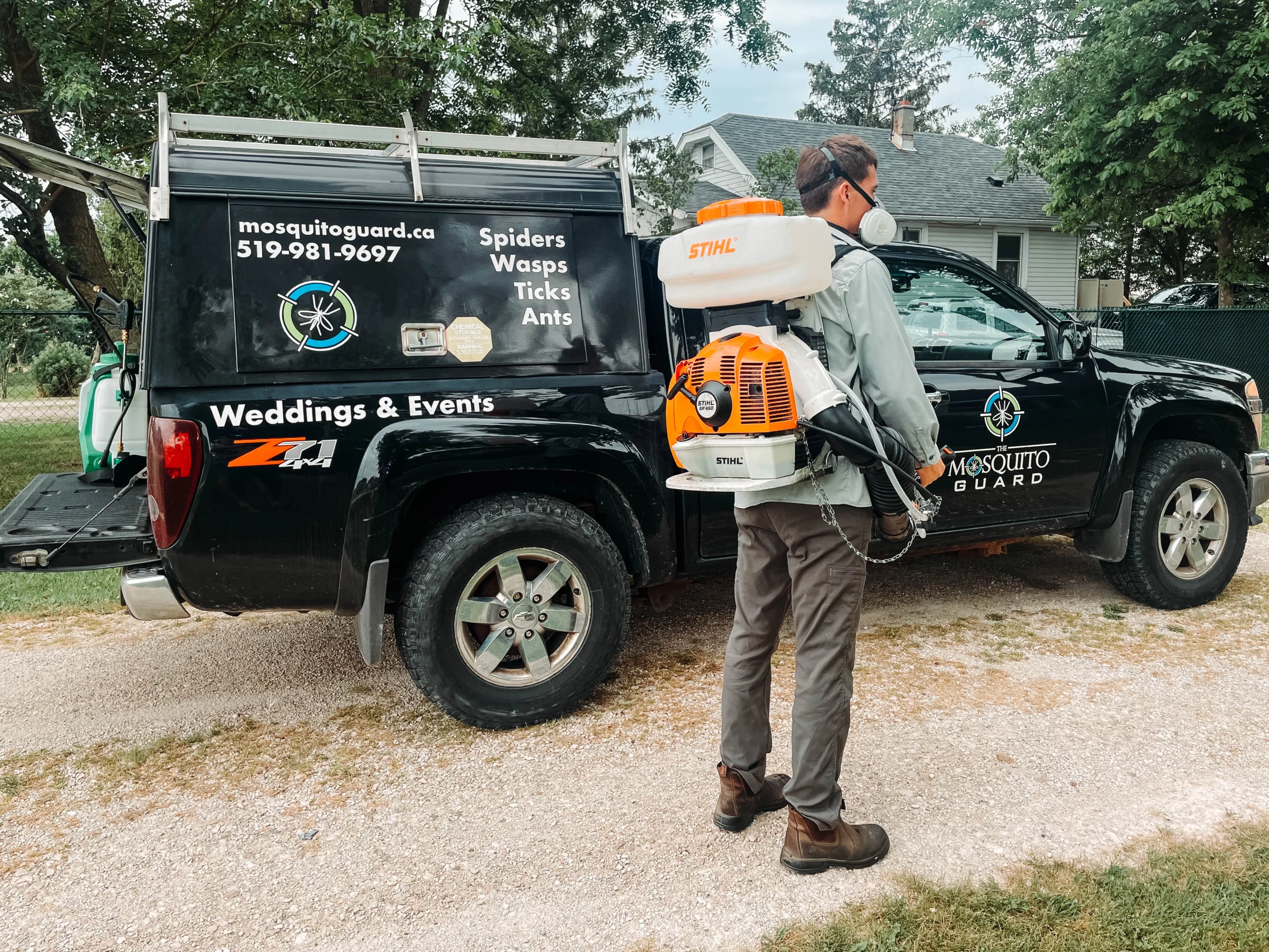 A Mosquito Guard technician wearing protective gear and a STIHL SR 450 backpack sprayer stands beside a black Mosquito Guard service truck. The truck displays the company’s contact information and pest control services, including spiders, wasps, ticks, and ants. A residential home and trees are visible in the background.