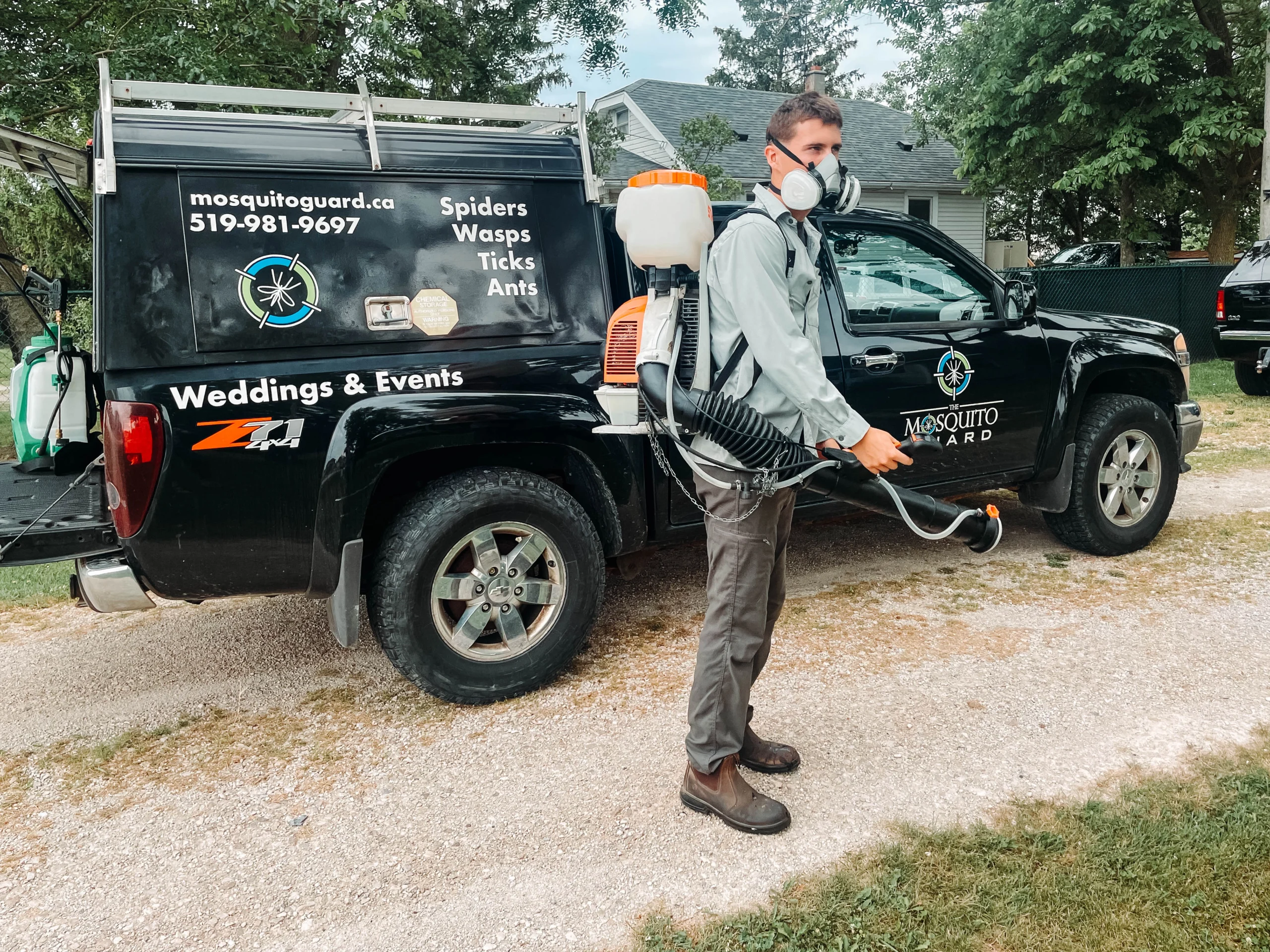Mosquito Guard technician wearing a respirator and operating a backpack sprayer, standing beside a branded black service truck that lists pest control services for spiders, wasps, ticks, and ants, with the slogan “Weddings & Events.”